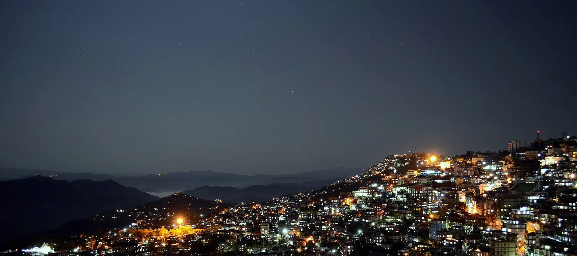 Aizawl at night — thousands of lights tracing the city down its ridgeline, with moonlit ranges and a thin band of valley mist fading into the distance