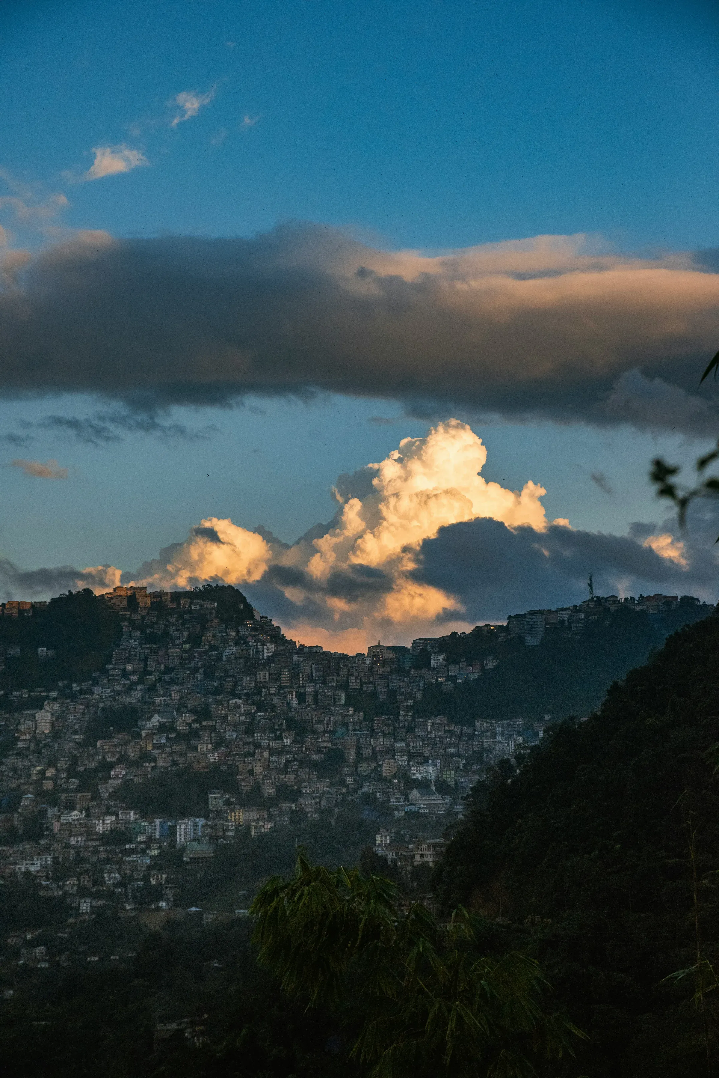 Aizawl's houses stacked up steep forested ridges at dusk, a sunlit cumulus cloud rising behind the hilltop skyline under a deep blue sky