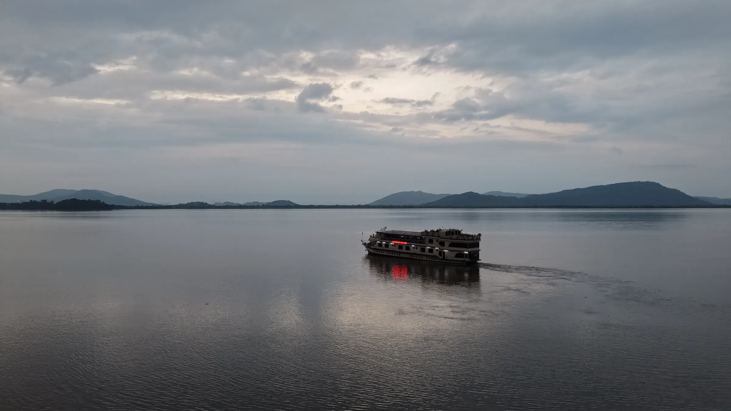 The Alfresco Grand cruise vessel small on the wide grey Brahmaputra in flood season, a line of hills along the far bank under an overcast sky