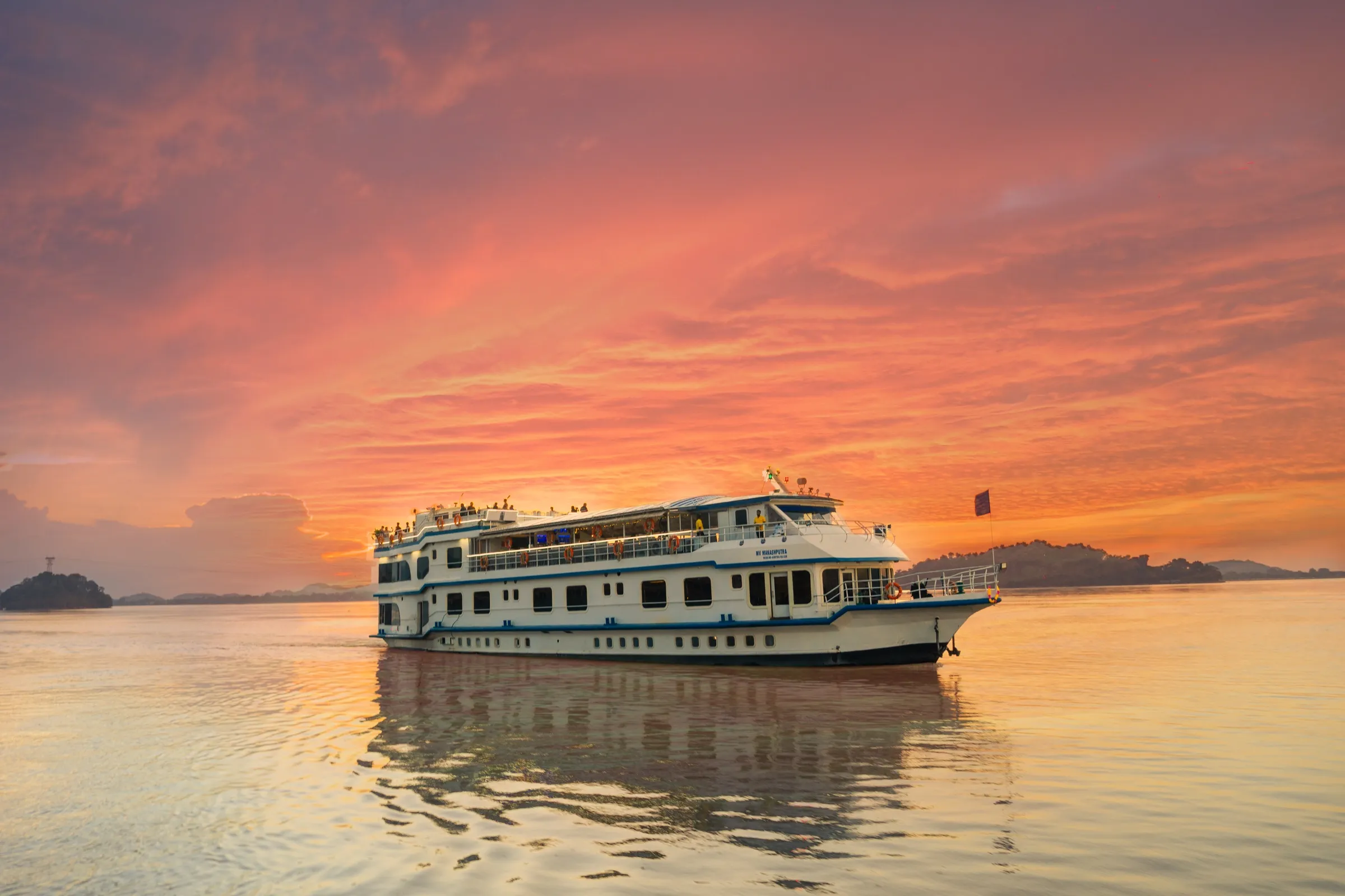 Alfresco Grand on the Brahmaputra under a blazing pink and orange sunset, guests on the open upper deck
