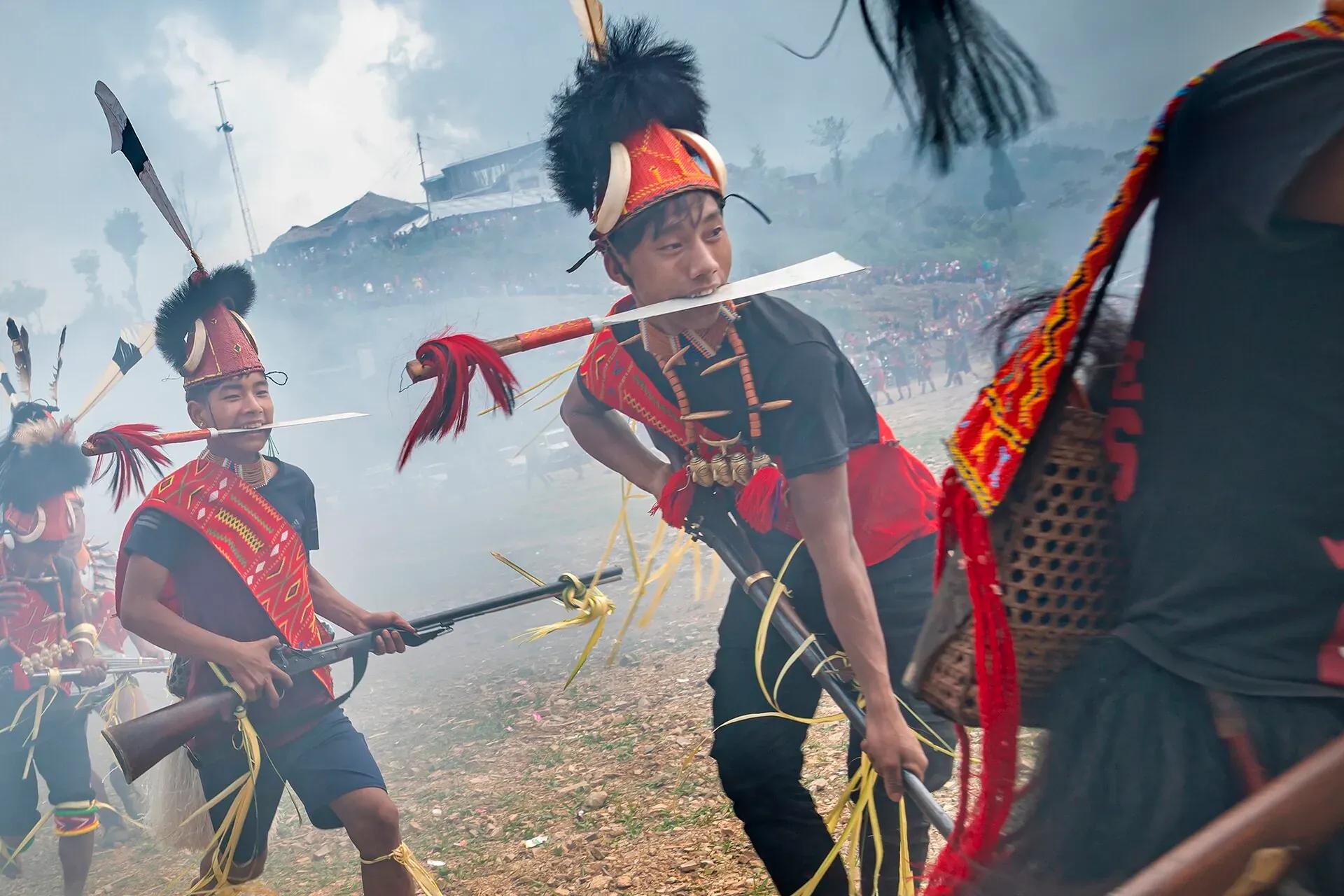 Young Konyak men mid-dance at Aoleang in Mon, wearing feathered headgear and red beadwork over everyday clothes, moving through smoke haze on a crowded festival ground
