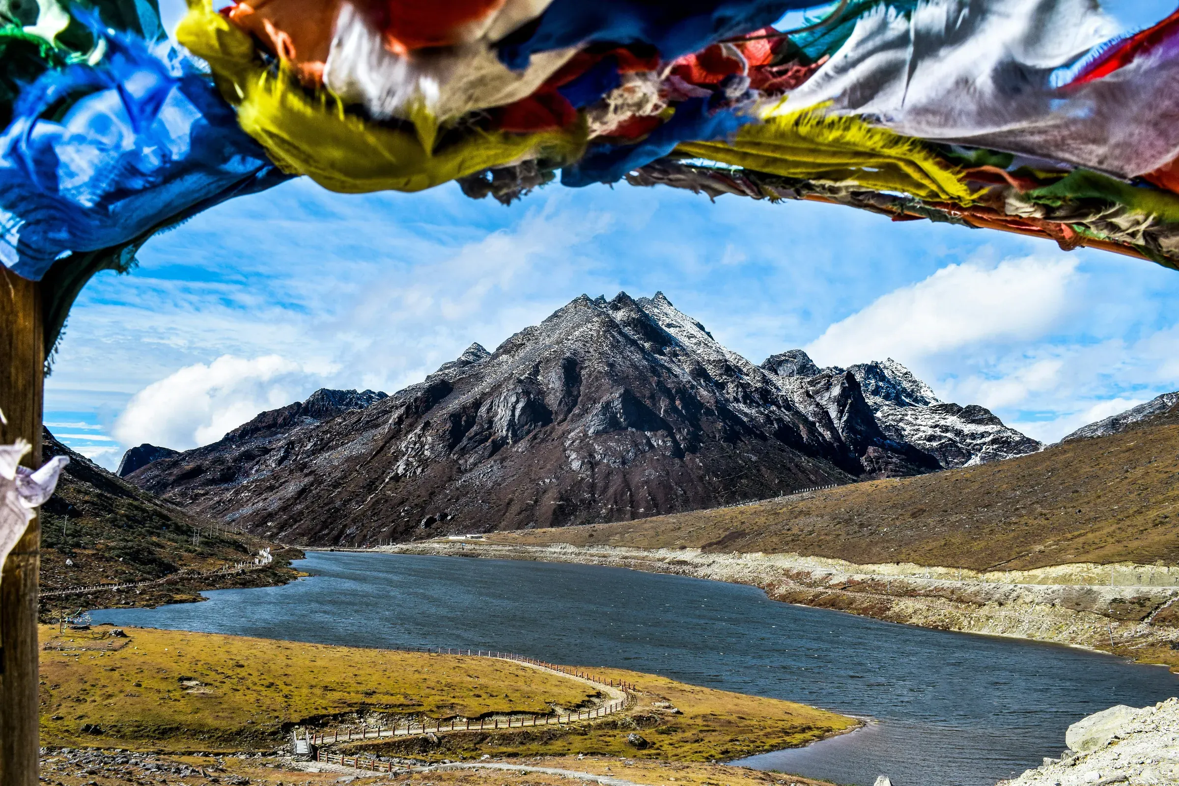 Snow-dusted Himalayan peaks above a high-altitude lake in Arunachal Pradesh, framed by an arch of colourful prayer flags