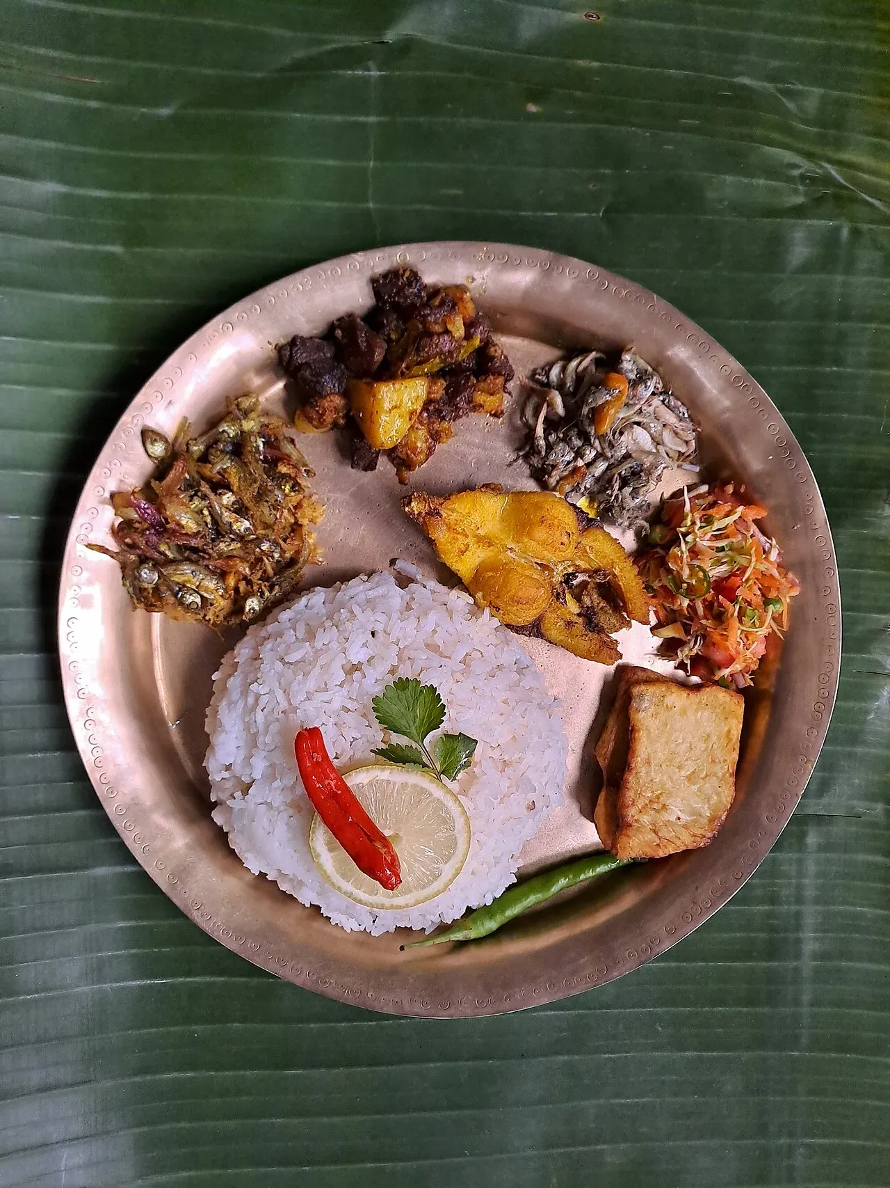 Assamese thali on a bell-metal plate over a banana leaf — a mound of rice with lemon and chilli, fried river fish, meat curry, fritters and fresh salad