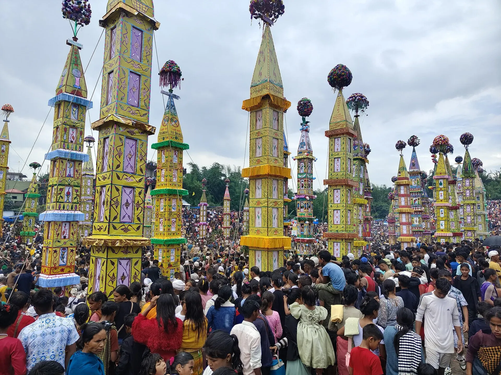 Tall, brightly painted tower structures raised above a dense festival crowd at Behdienkhlam in Jowai, under a heavy monsoon sky