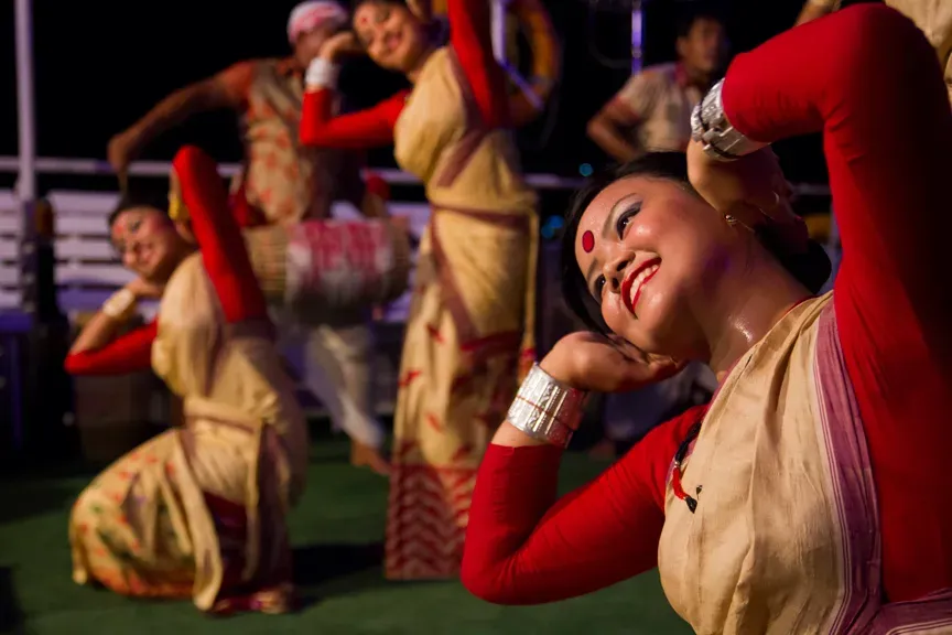 Bihu dancers in cream and red traditional dress performing on the deck of Alfresco Grand, a smiling dancer in the foreground and a dhol drummer behind