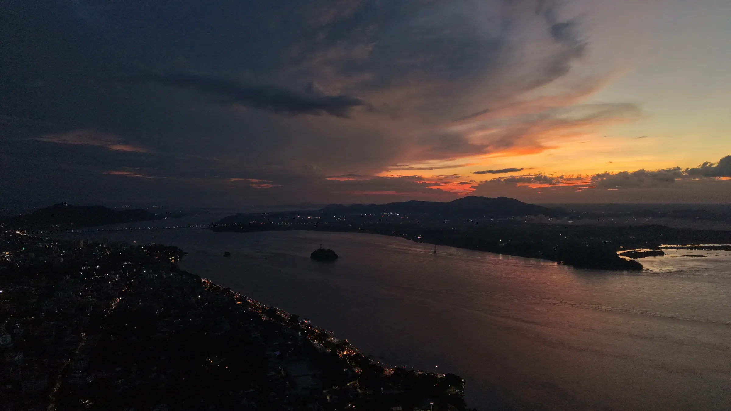 Aerial view of the Brahmaputra at Guwahati at dusk, city lights along the bank and an orange sunset behind the hills