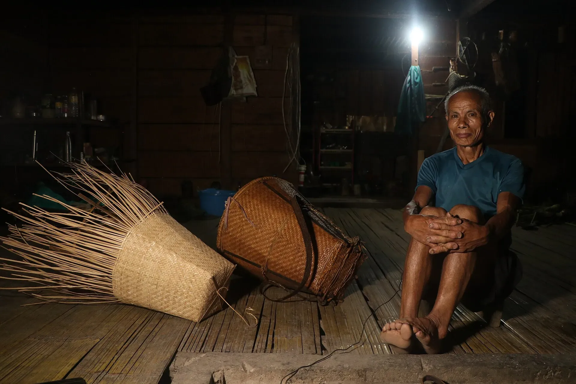 An artisan seated on the bamboo floor of a dim wooden house beside two woven carrying baskets, one still in progress with long cane strips fanning out unfinished