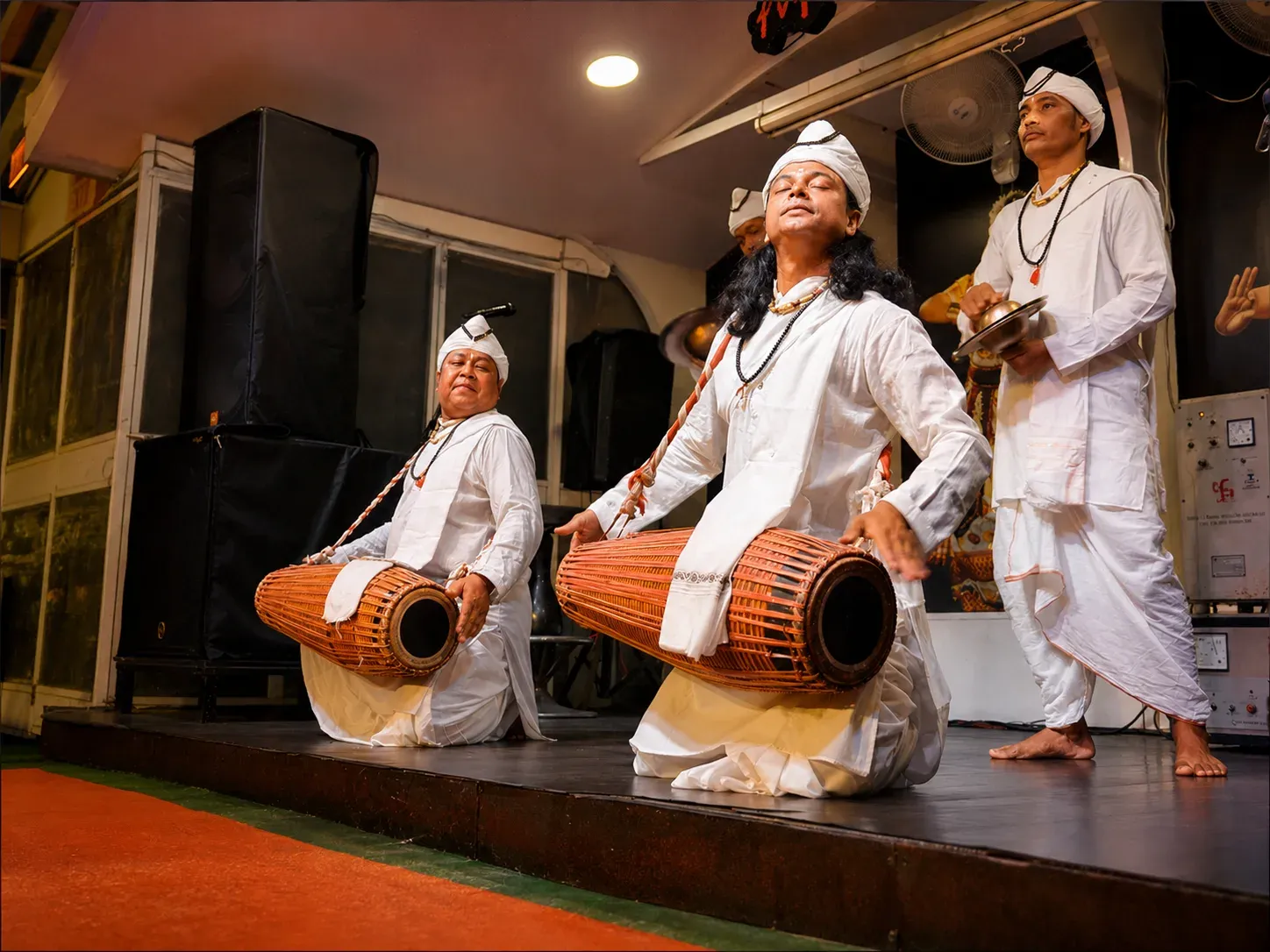 Gayan Bayan performers in white robes and turbans playing khol drums and cymbals on the Alfresco Grand stage