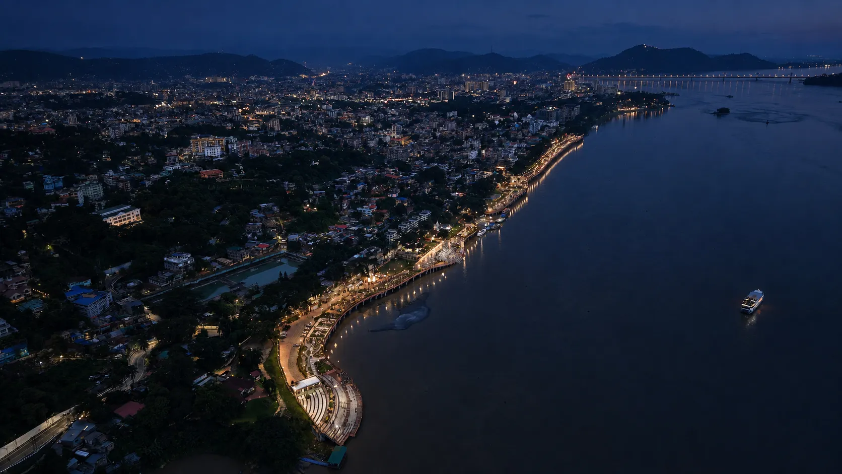 Guwahati's riverfront lights at night from above, with a lit cruise vessel on the dark Brahmaputra
