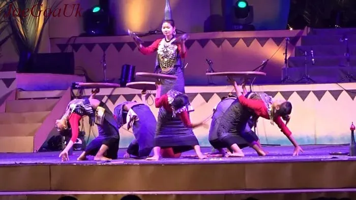 A Hojagiri performance on a lit stage — a row of Reang dancers in dark skirts and red blouses bending deeply forward, two balancing round tabletops, while a lead dancer stands upright behind them