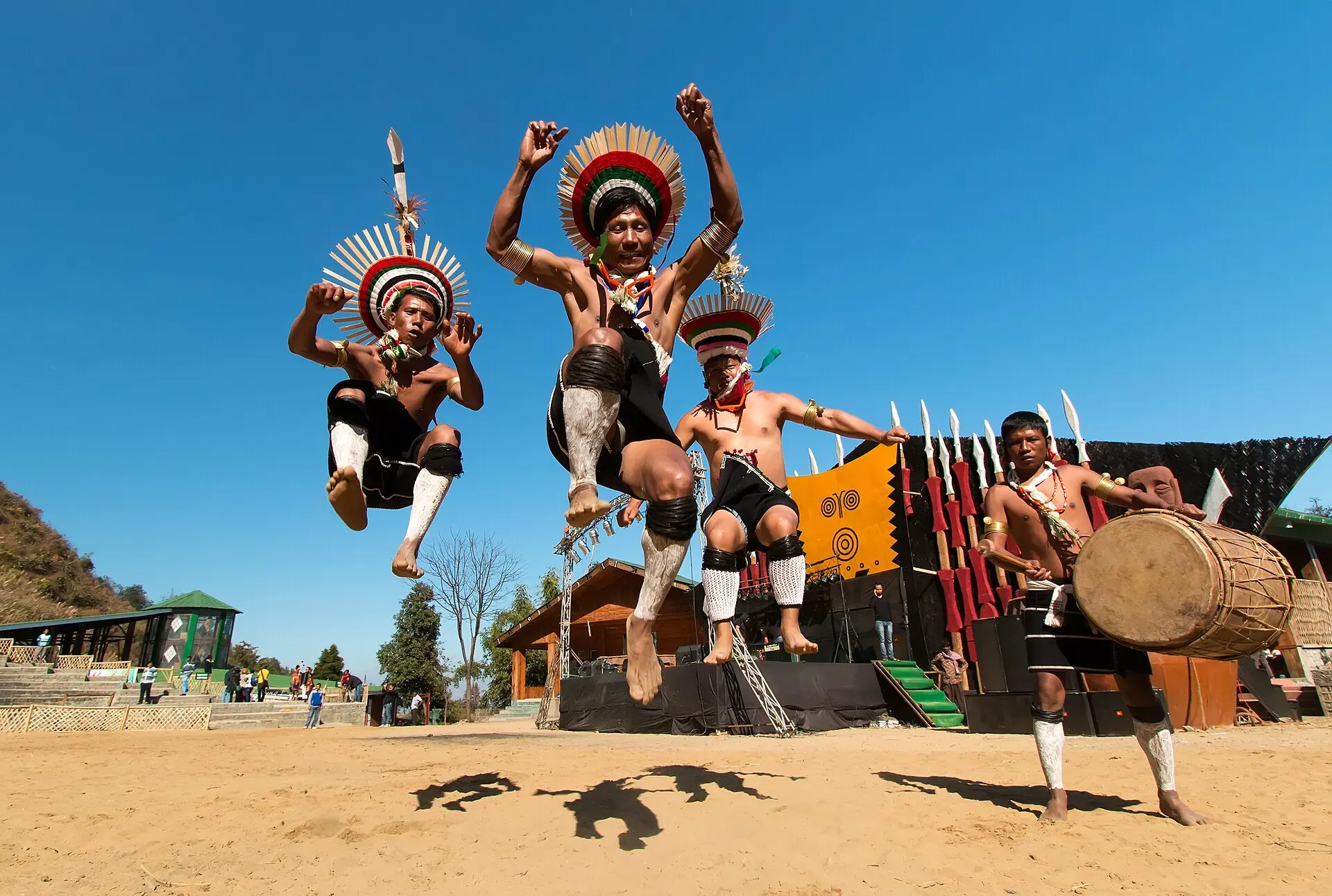 Dancers leaping high in unison at the Hornbill Festival, wearing flat circular cane headdresses and white leggings, with a log-drummer beside them and a morung-style stage behind