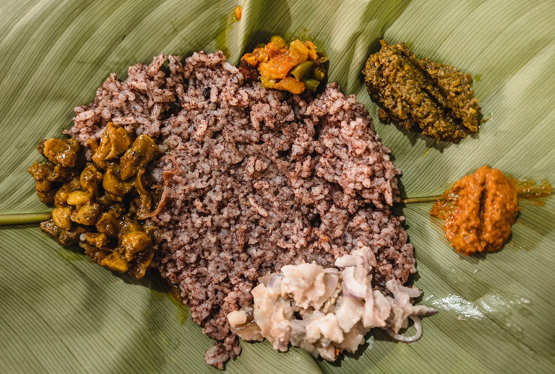 Khasi meal served on a banana leaf — reddish-brown jadoh rice at the centre, ringed by pork, leafy greens, chutneys and an onion salad