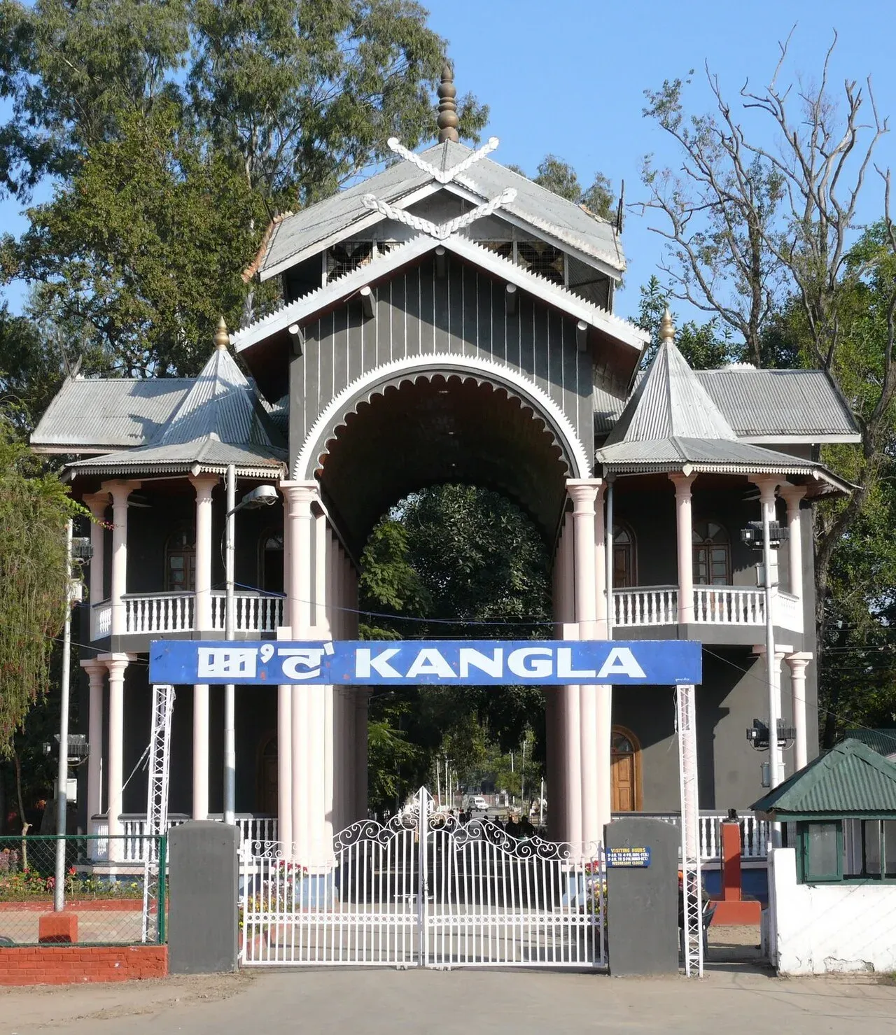 The Kangla Gate in Imphal — a pale arched gateway with tiered grey roofs and slender columns, a blue KANGLA sign above white entrance gates, tall trees behind