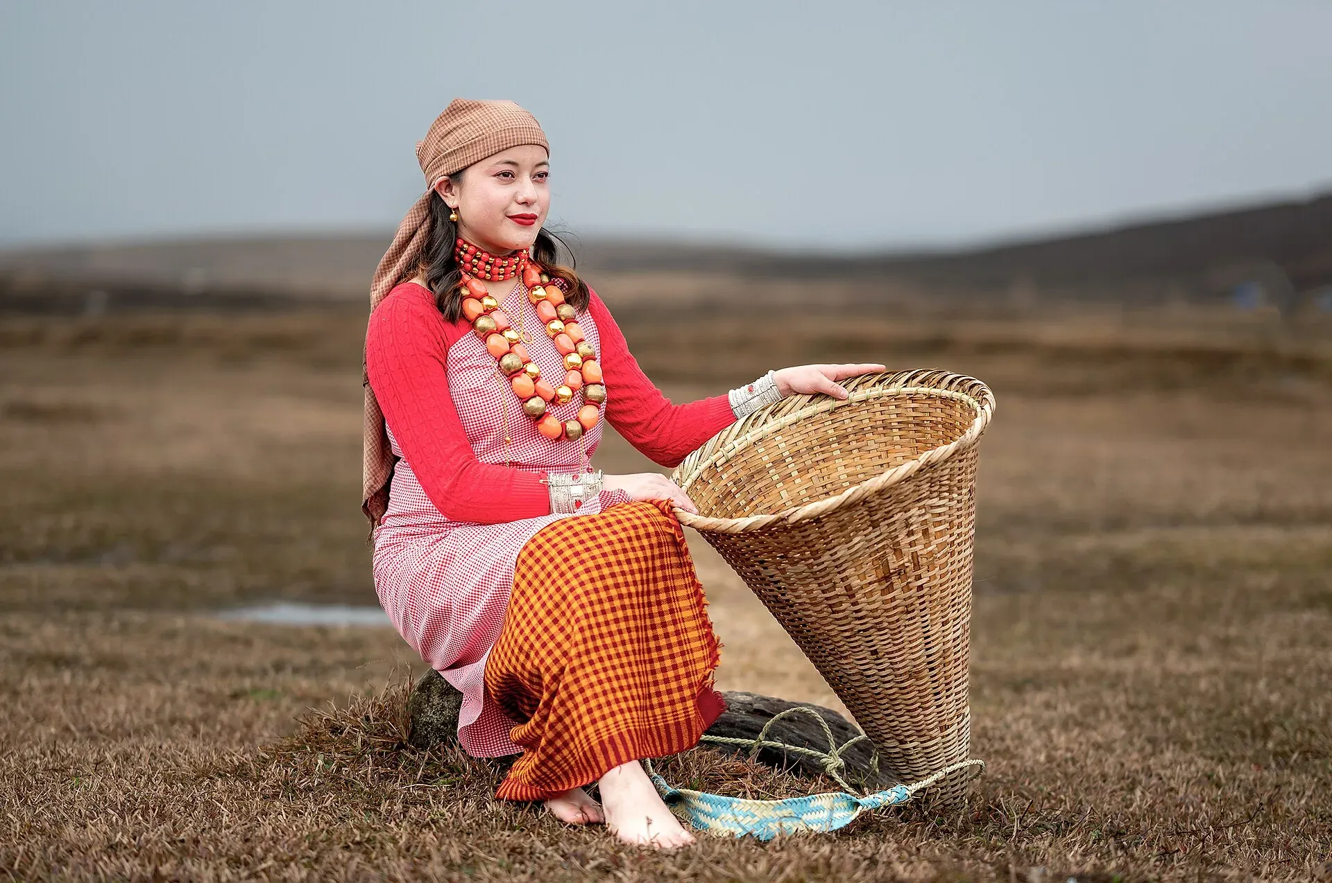 Woman in traditional Khasi dress with coral and gold beads seated in open grassland beside a large conical woven bamboo khoh basket