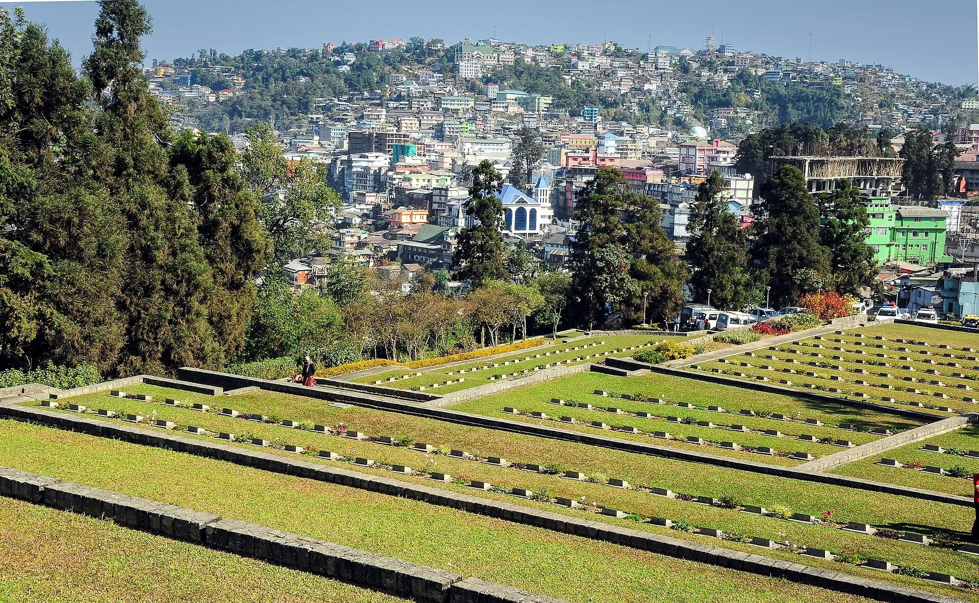 Terraced lawns of the Kohima War Cemetery with rows of low stone grave markers, the hillside houses of Kohima rising behind