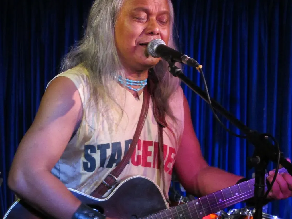 Lou Majaw singing into a microphone with an acoustic guitar, long silver hair falling over a sleeveless shirt, in front of a blue stage curtain