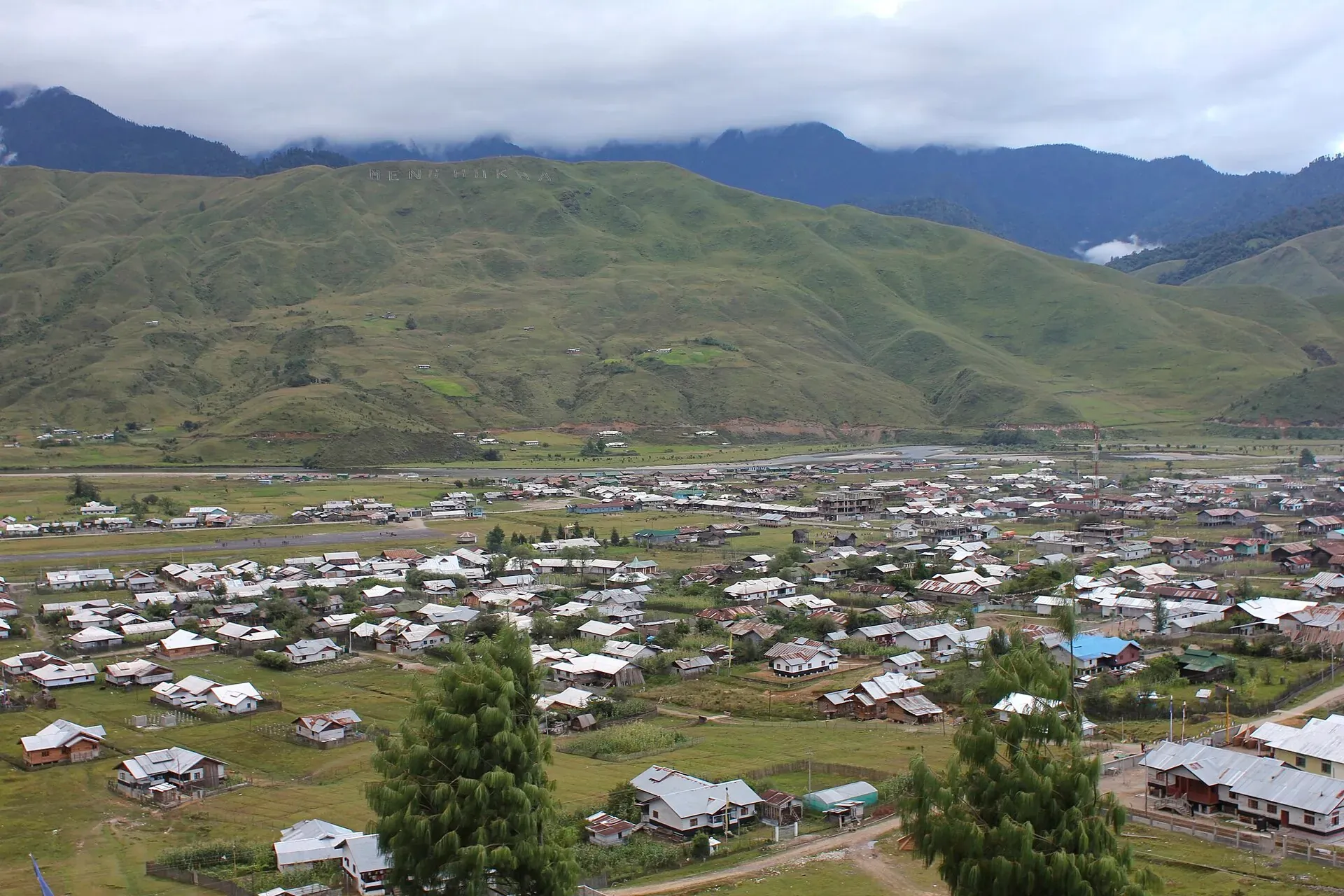 Mechuka town spread across a wide, flat valley floor — scattered tin-roofed houses on open grassland below bare green hills with clouds resting on the ridgelines