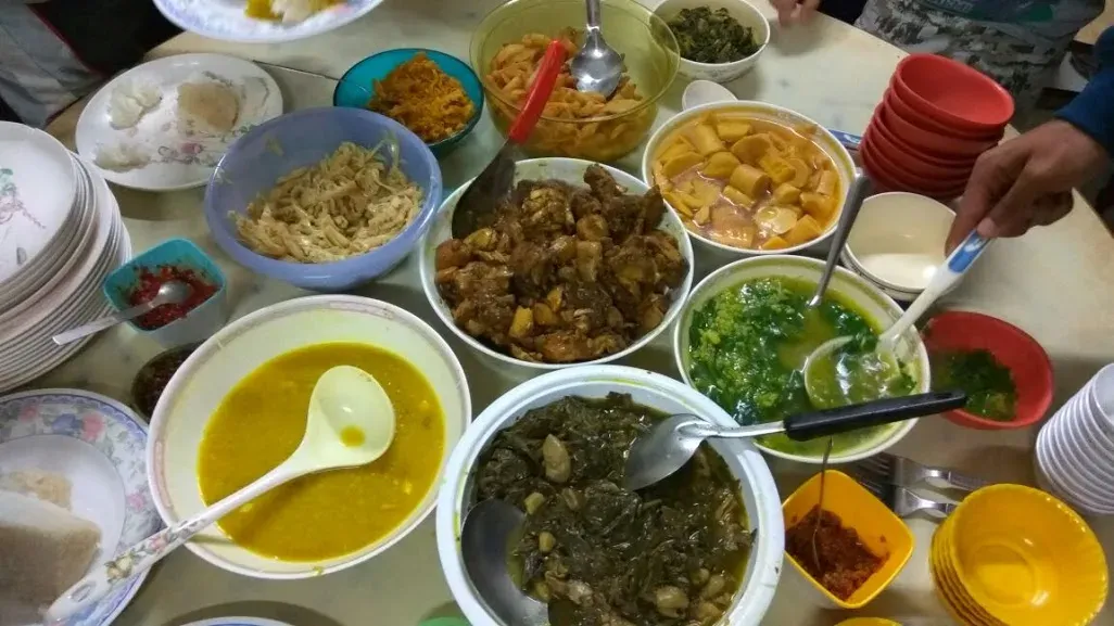A Mizo home spread on a table — bowls of dal, stewed greens with pork, a green leafy broth, boiled vegetables, bamboo shoot, chutneys and plates of rice being served
