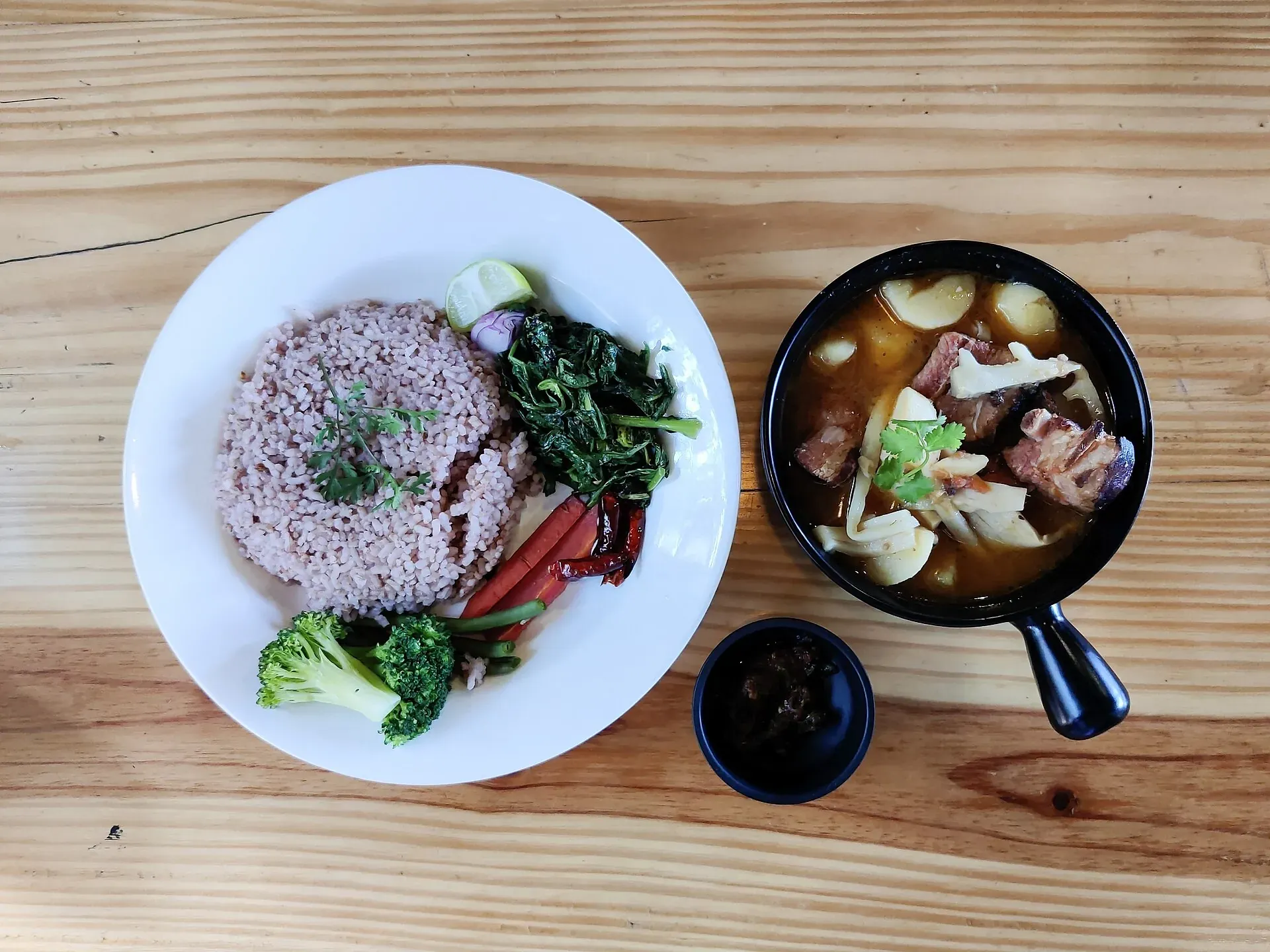 Naga meal on a wooden table — a bowl of smoked pork curry with bamboo shoot, a plate of brown rice with sautéed greens, broccoli and chillies, and a small dish of dark chutney