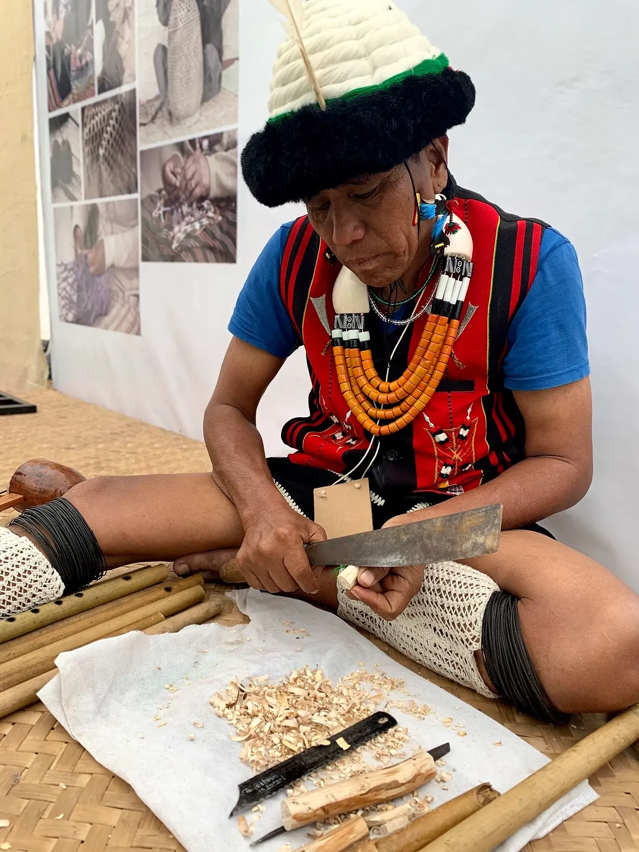 A Naga artisan in ceremonial dress — feathered cap, orange bead necklaces and woven leggings — carving a piece of wood with a dao at a craft demonstration, shavings and bamboo flutes on the mat before him