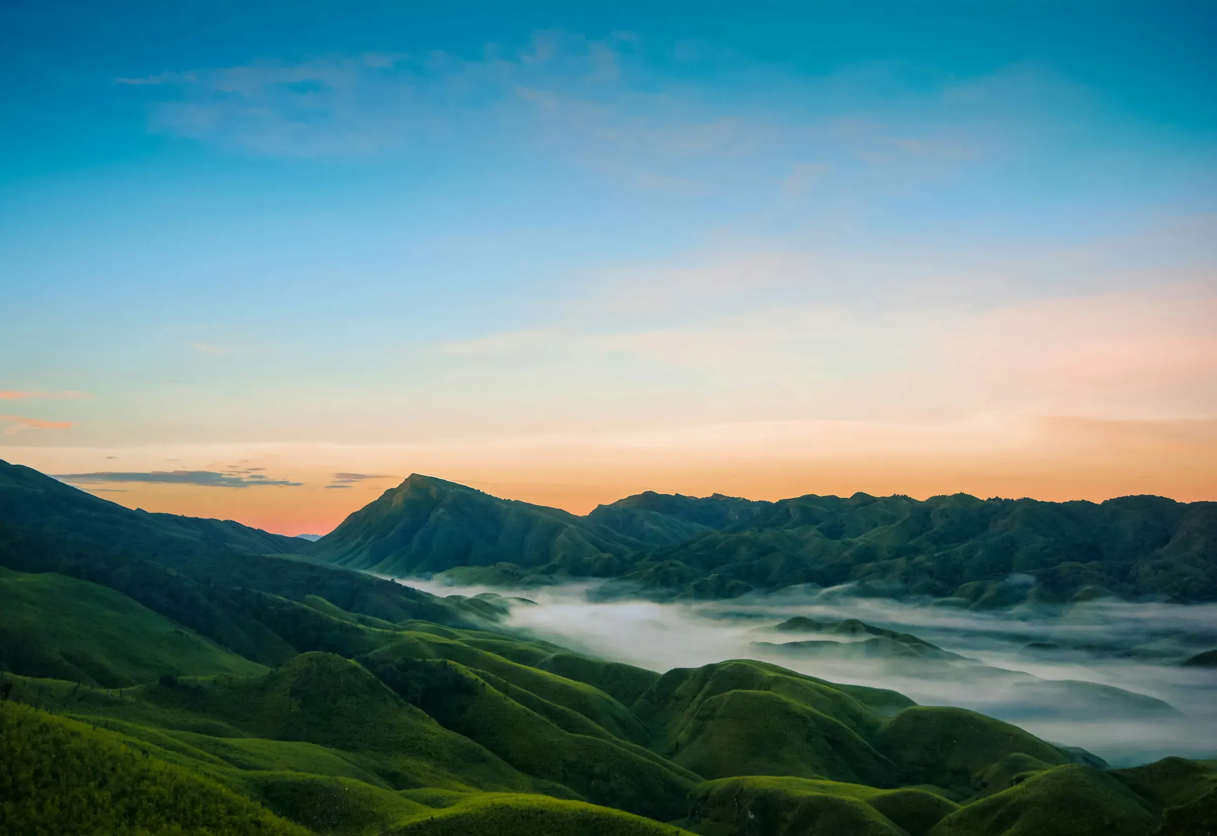Rolling green hills of Nagaland at dawn, mist pooled in the valley under a sky turning from orange to blue