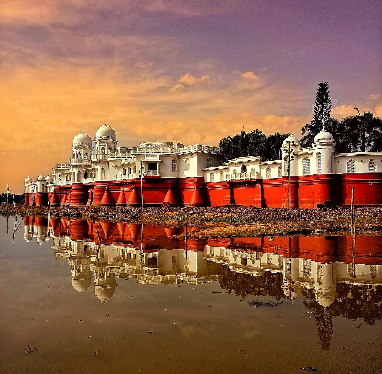 Neermahal palace stretched along the edge of Rudrasagar Lake at sunset — white domes and pavilions above red-banded walls, the whole facade mirrored in the still water
