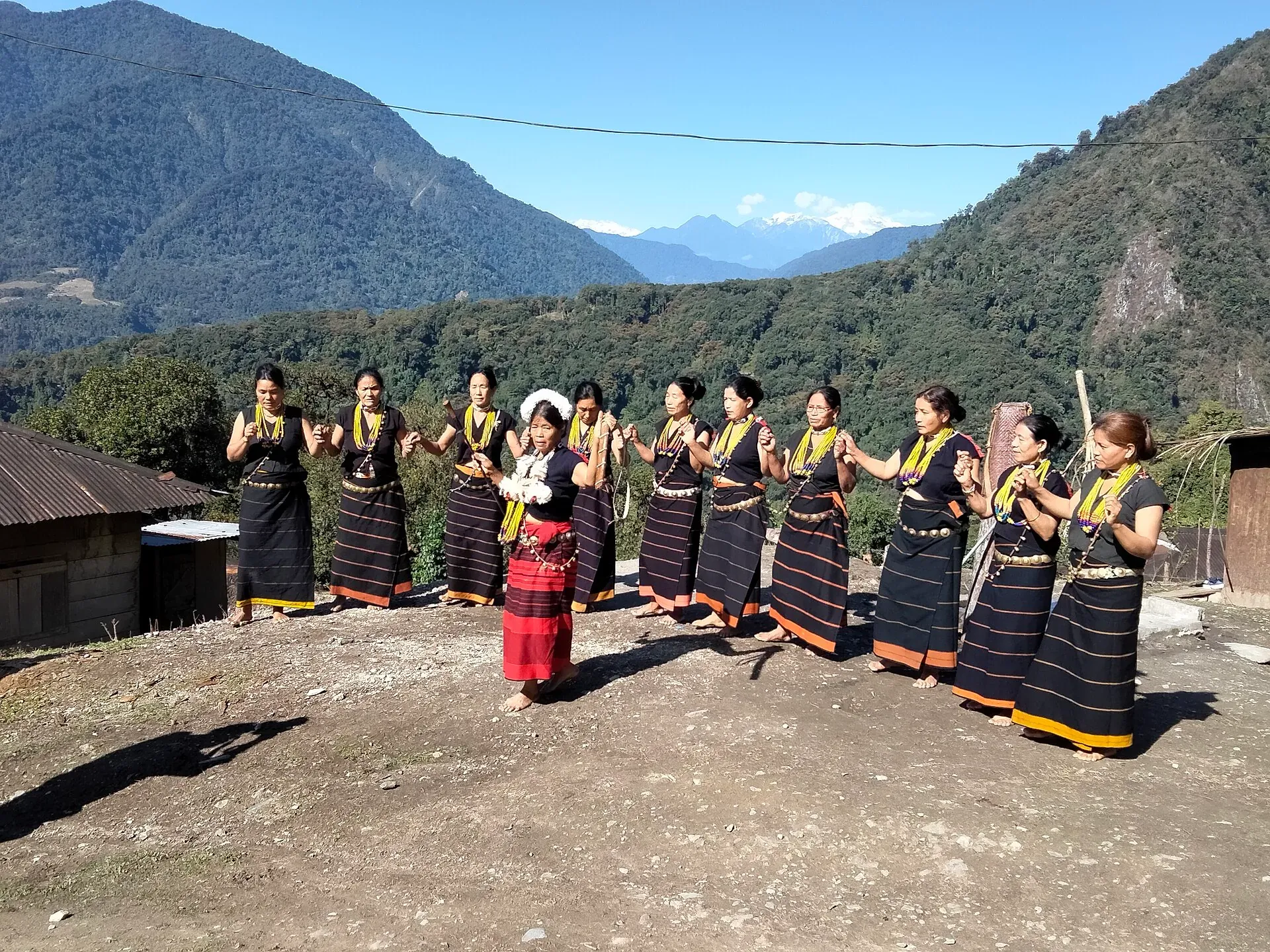 A line of Adi women in black-and-orange striped wraps and yellow bead necklaces dancing hand in hand on a village clearing, a lead performer in red at the centre and forested mountains behind