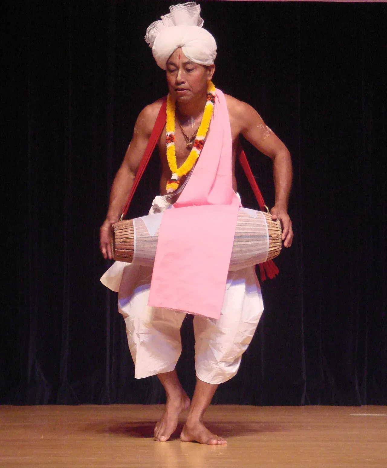 A Pung Cholom performer in white dhoti, white turban and marigold garland caught mid-step on one foot while playing the barrel-shaped pung drum slung from his shoulder