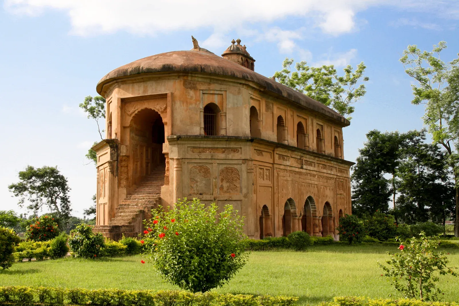 Rang Ghar at Sivasagar — a two-storey Ahom-era amphitheatre in ochre brick with an oval roof, arched openings and a staircase, set in lawns with flowering shrubs