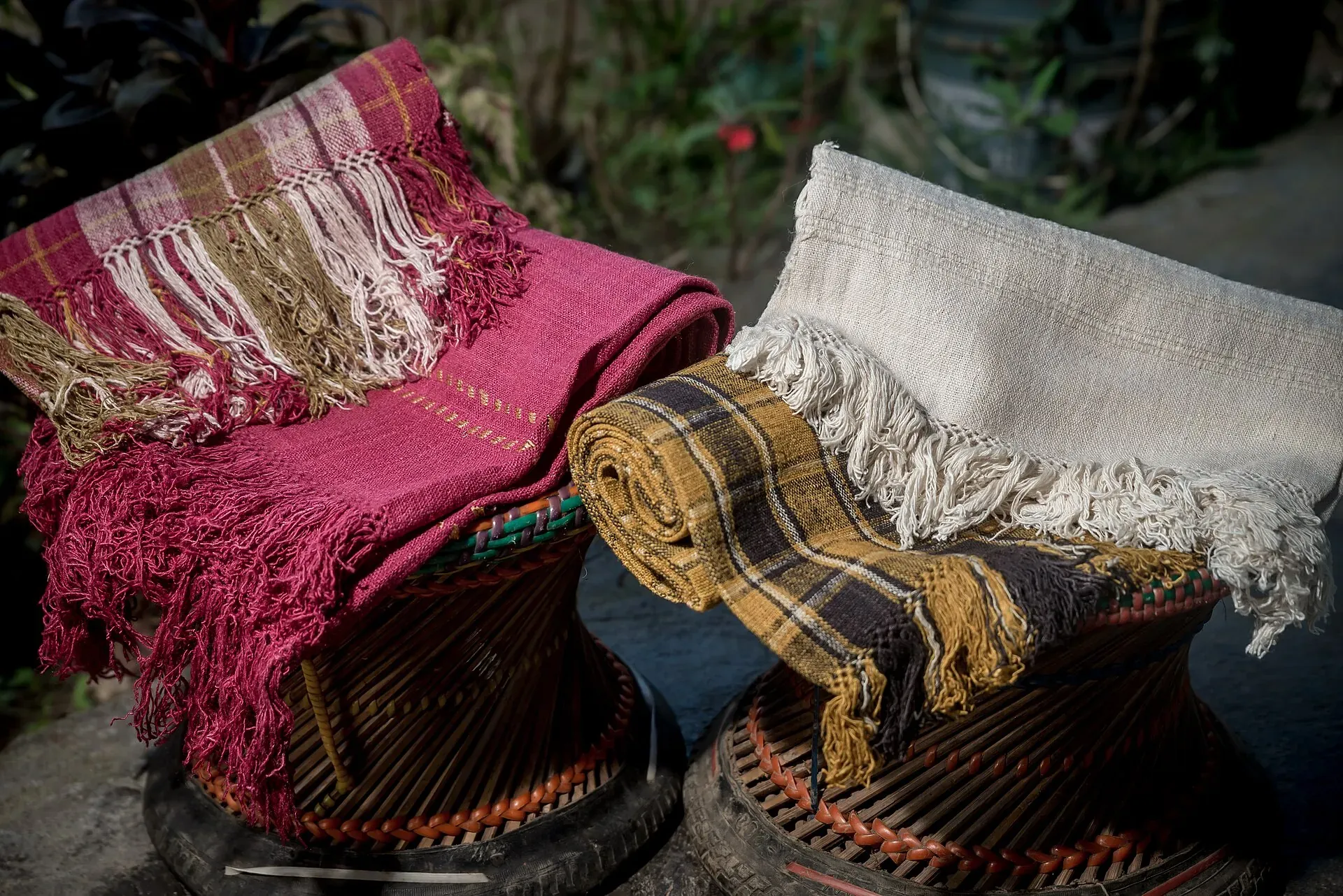Handwoven ryndia eri-silk shawls — one madder red with fringed panels, one mustard and black check, one undyed cream — folded over cane stools in sunlight