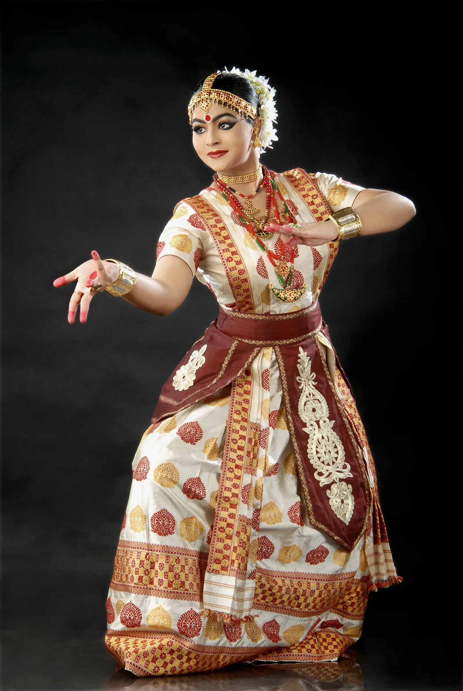 Sattriya dancer in a cream and red pat silk costume with traditional Assamese jewellery, holding a narrative hand gesture against a dark background