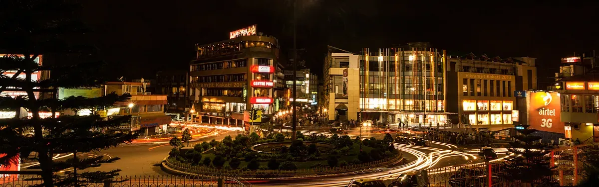 Night panorama of a busy Shillong commercial junction — lit shopfronts and signboards around a roundabout, with light trails from circling traffic