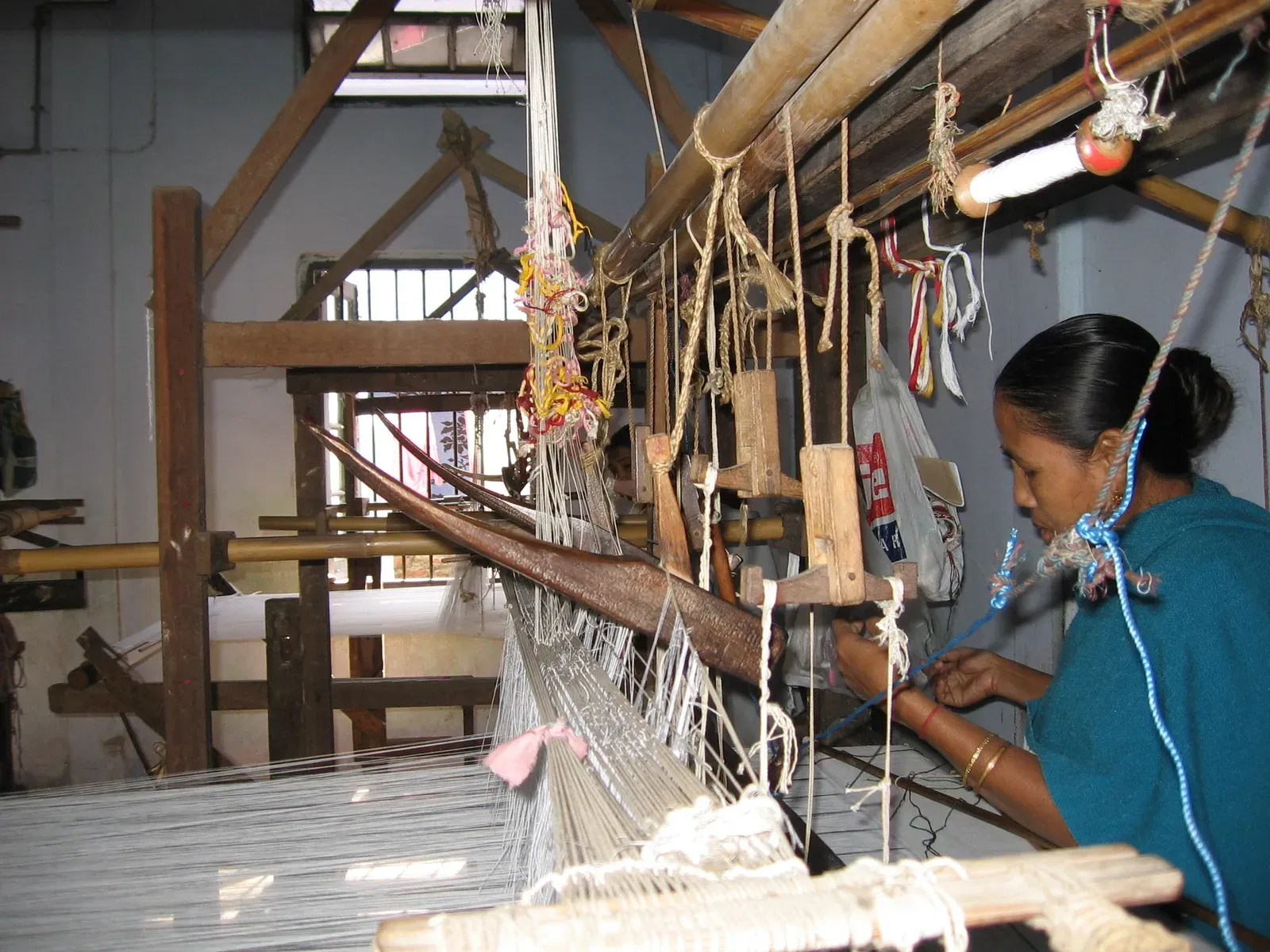 Weaver working at a large wooden handloom in Sualkuchi, drawing pale silk warp threads strung across the frame