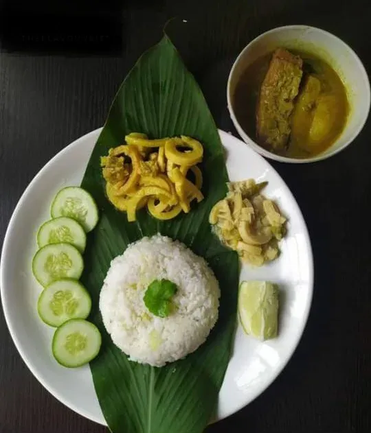 A Tripuri meal served on a banana leaf — a mound of rice with sliced cucumber, bamboo shoot curry, boiled vegetables and lime, and a bowl of fish in a green herb gravy alongside