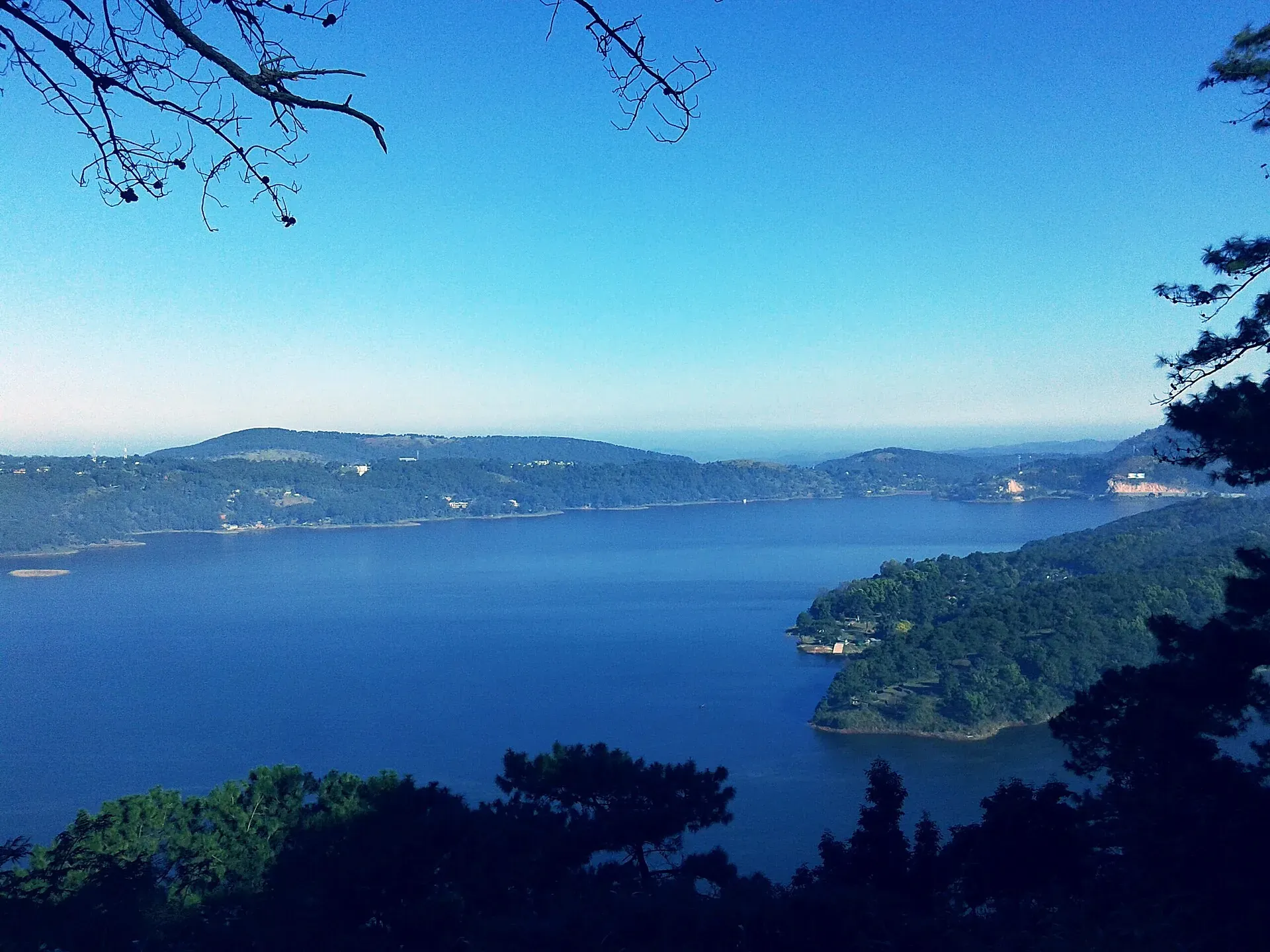 Umiam Lake from a hillside viewpoint on the Guwahati–Shillong road — broad blue water reaching between pine-covered ridges under a clear sky