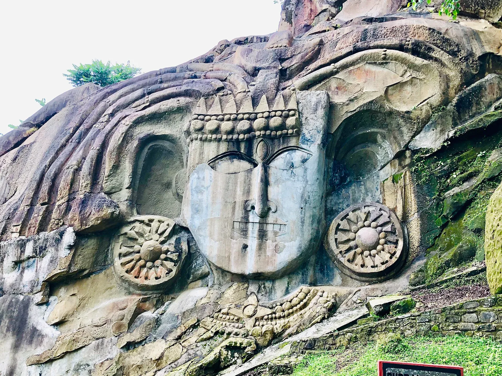 The colossal rock-cut face of Shiva at Unakoti, wearing a carved crown and flanked by two large rosette discs, emerging from the layered rock of the forested hillside