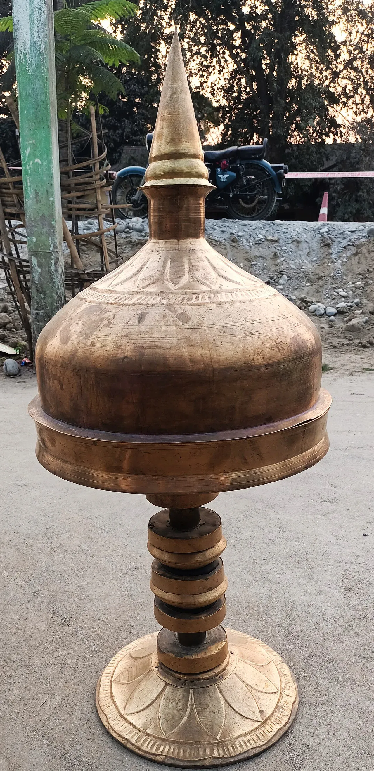 Tall bell-metal xorai with a domed, spired lid standing on a pedestal outside a workshop