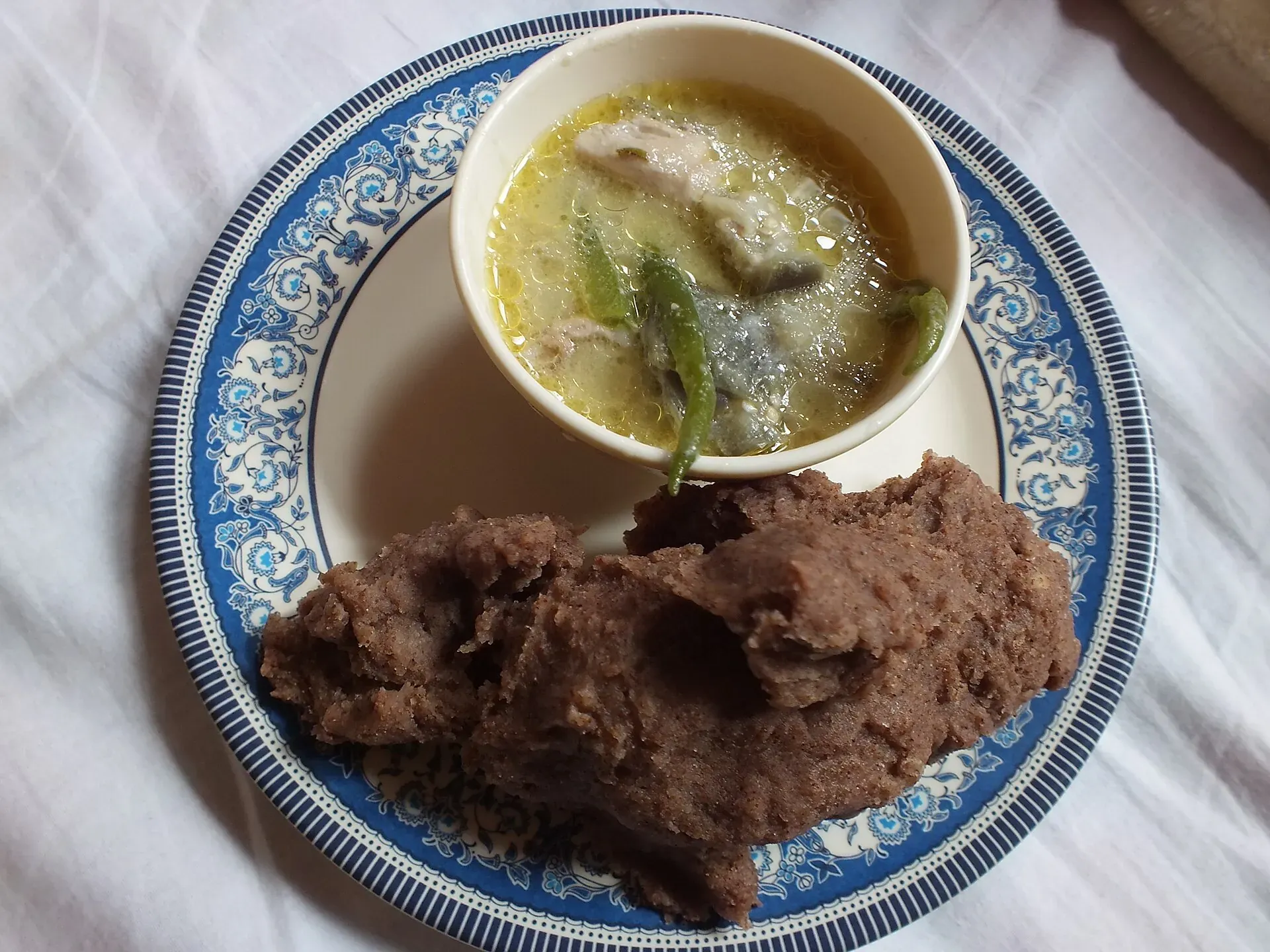 A serving of zan — a dark millet-flour porridge — on a blue-patterned plate beside a small bowl of broth with meat, greens and green chillies